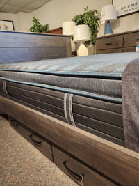 A close-up of the Beauty-rest Isla PT Firm Mattress on a wooden bed frame, with lamps and plants in the background.
