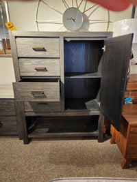 The Magnussen B5990 Plum Creek Door Chest in Ravenwood Black has 3 drawers, open shelves, and a clock behind it.