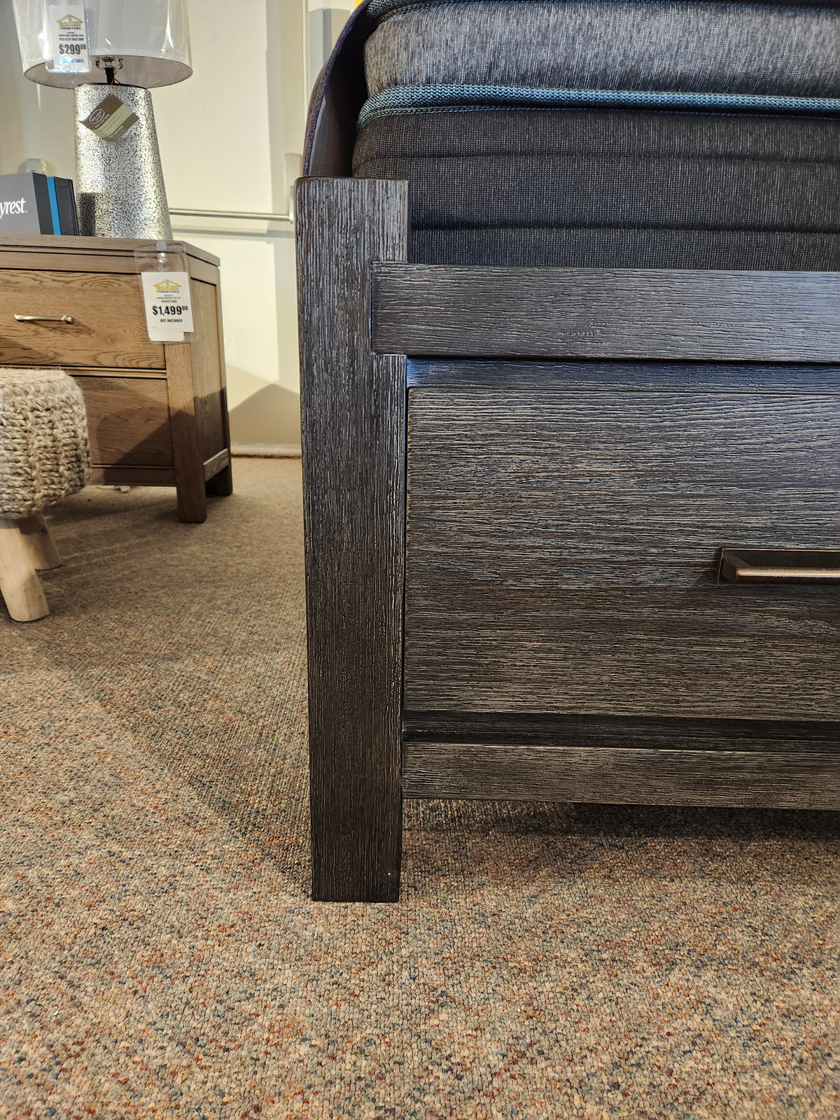 Close-up of the Magnussen Plum Creek B5990 Queen Storage Bed in Ravenwood Black, with drawer, next to a nightstand.