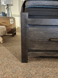 Close-up of the Magnussen Plum Creek B5990 Queen Storage Bed in Ravenwood Black, with drawer, next to a nightstand.