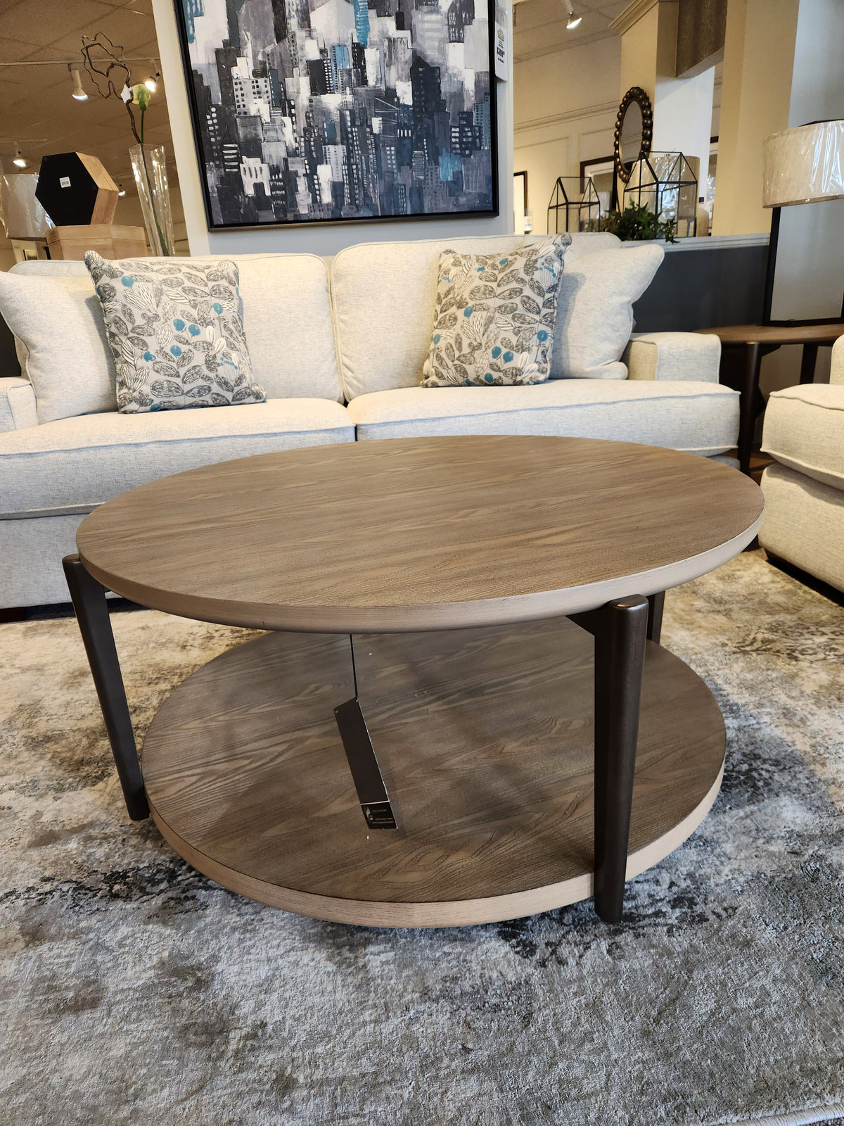 The Ashley Dyonton Round Cocktail Table features a wood-tone finish and lower shelf for an industrial, modern showroom look.