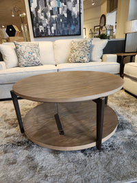 The Ashley Dyonton Round Cocktail Table features a wood-tone finish and lower shelf for an industrial, modern showroom look.
