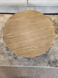 The Ashley Dyonton Round Cocktail Table with wood-tone finish sits on a rug before a light-colored upholstered sofa.