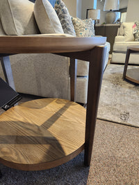 The Ashley Dyonton Round End Table with wood-tone finish is displayed beside a gray sofa with patterned pillows.