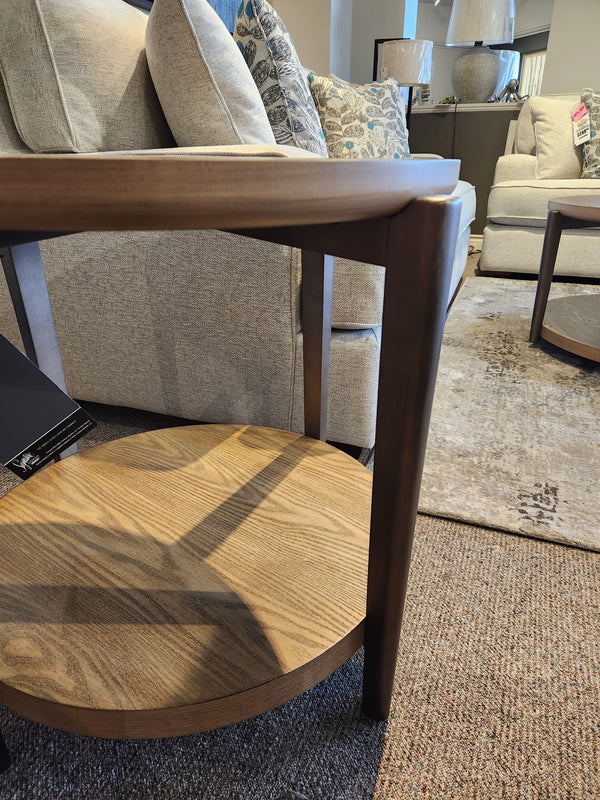 The Ashley Dyonton Round End Table with wood-tone finish is displayed beside a gray sofa with patterned pillows.