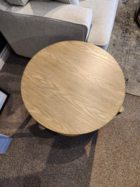 The Ashley Dyonton Round End Table with wood-tone finish brings contemporary style beside a beige sofa on brown carpet.