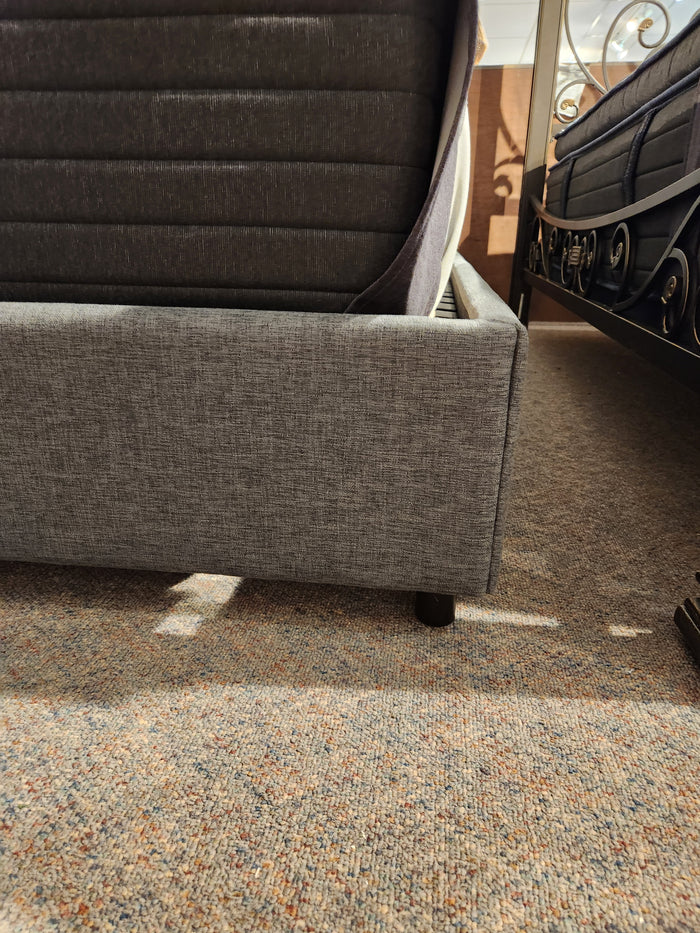 Donald Choi Alba Upholstered Bed Frame in grey with mattress sits next to a metal queen bed on a carpeted floor.