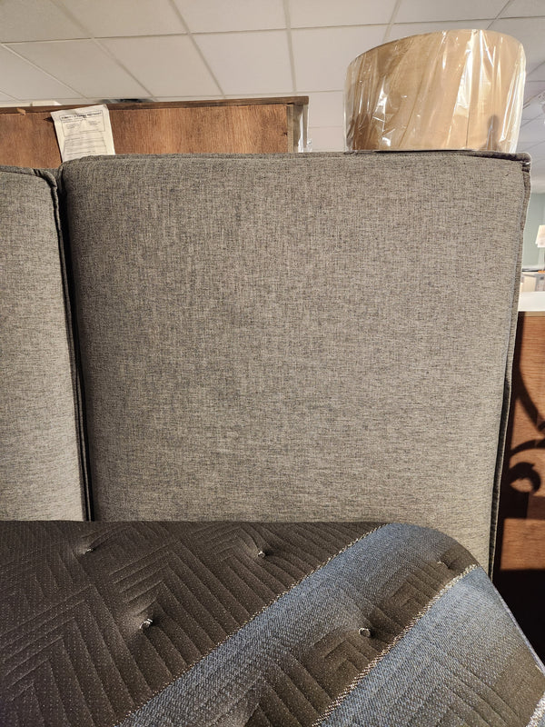 A gray Donald Choi Alba Upholstered Bed Frame and mattress are shown in a furniture store with a lamp behind.