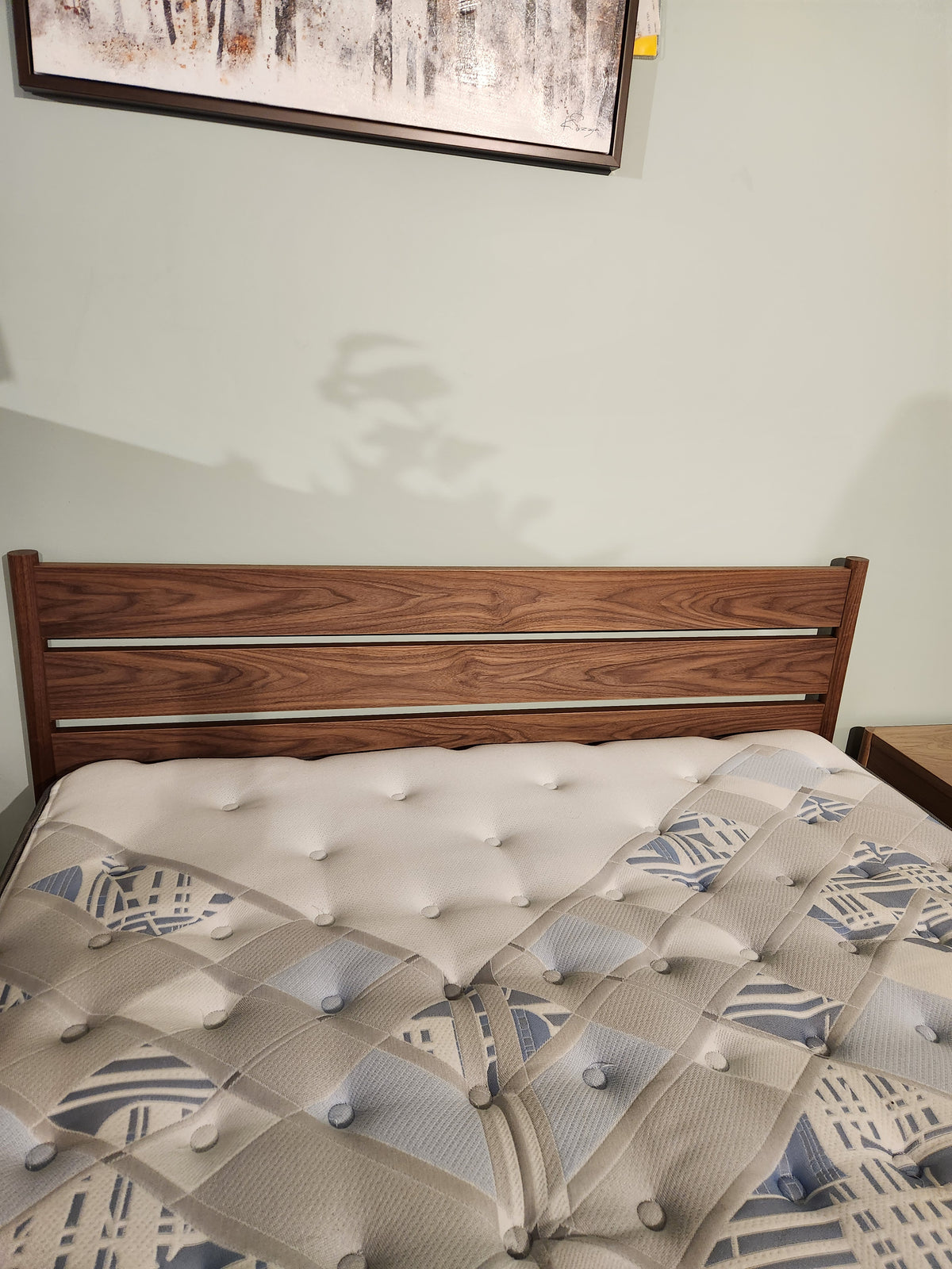 The Palliser EQ3 Monarch Queen Bedframe with a patterned mattress sits below framed art on a light-colored wall.