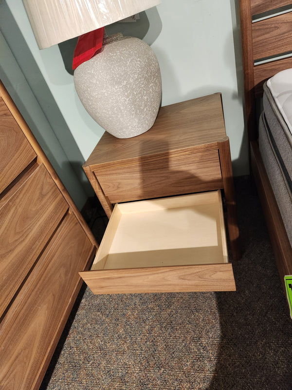 A Palliser EQ3 Monarch 2 Drawer Night Stand in walnut veneer sits by a bed and lamp, with one drawer open.