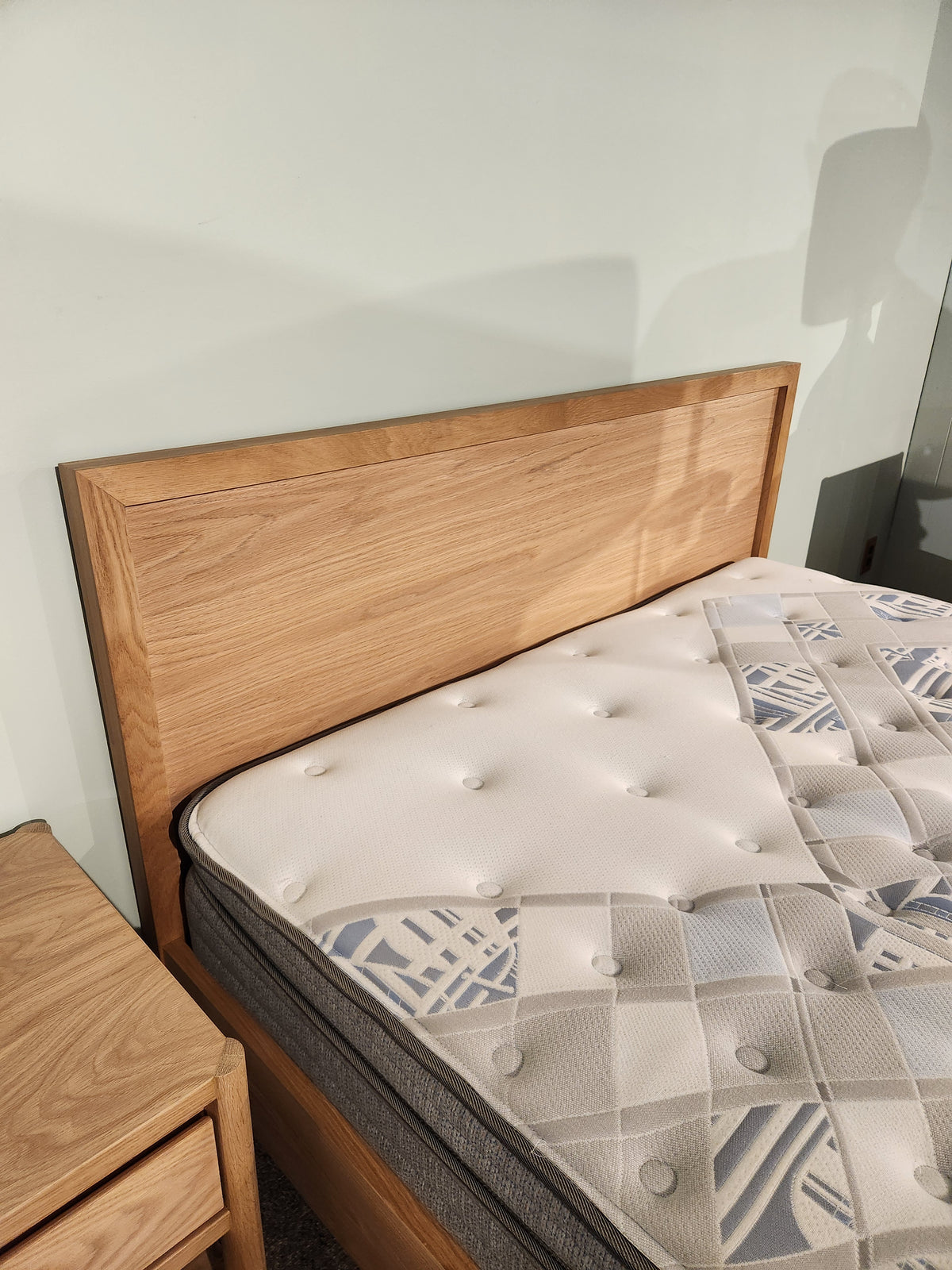A Palliser EQ3 Marcel Queen Bedframe with wood veneer stands by a matching nightstand against a light wall.