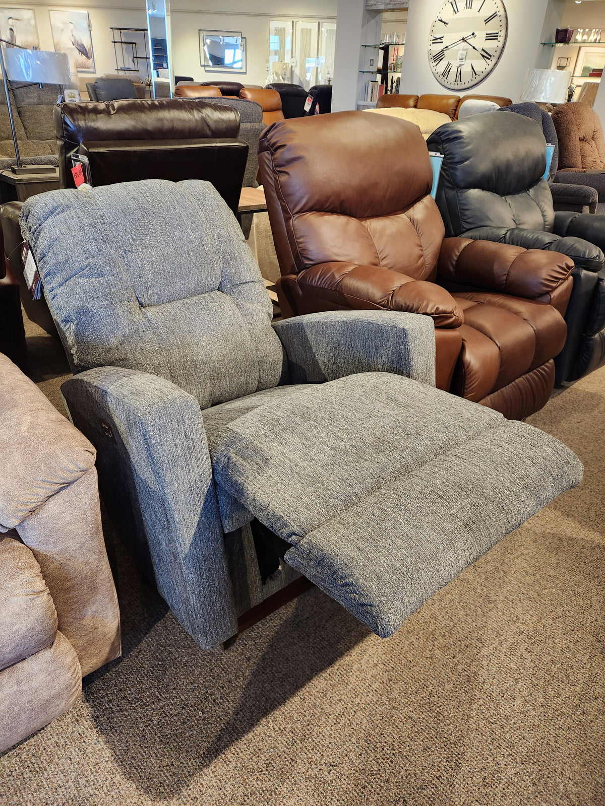 The La-z-boy 767 Casey Power Rocker Recliner in gray, with footrest extended, is displayed among other in-store styles.