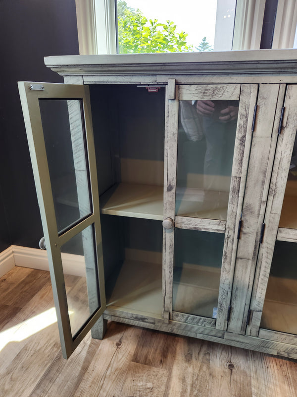 The Jofran Rustic Shores 4 Door Accent Cabinet stands open, showing empty shelves on a wooden floor beside a window.