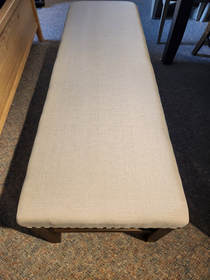 The Jofran Willow Creek Bench, with beige upholstery and wood legs, sits on carpet beside wooden furniture.