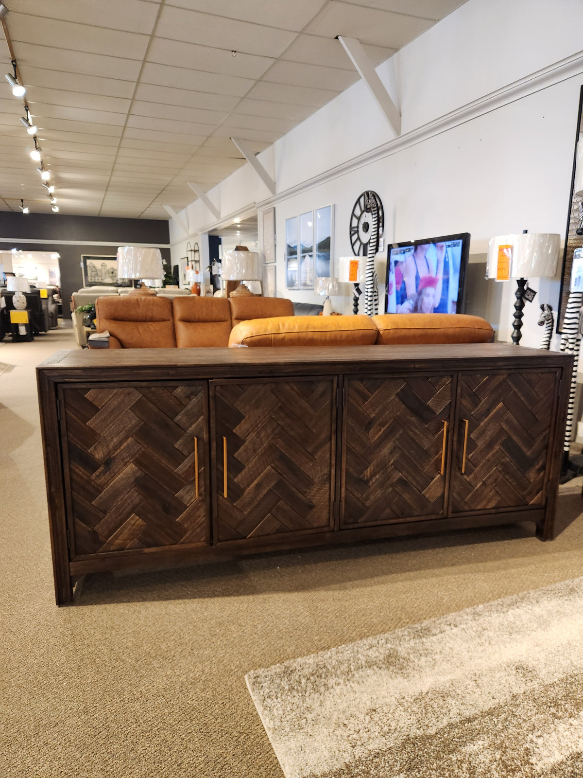 The Jofran Gramercy 4 Door Accent Cabinet features a chevron wood inlay and gold handles, shown in a modern showroom.