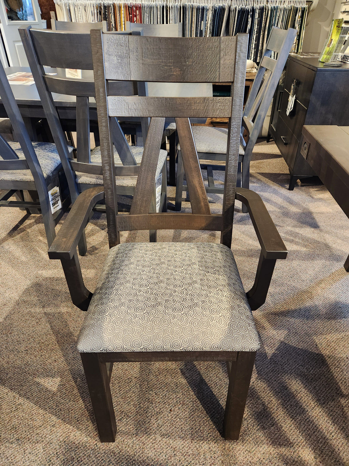 A dark wood armchair from Handstone's RA4284 Rafters Dining Package, with a patterned seat, shown among rustic chairs.