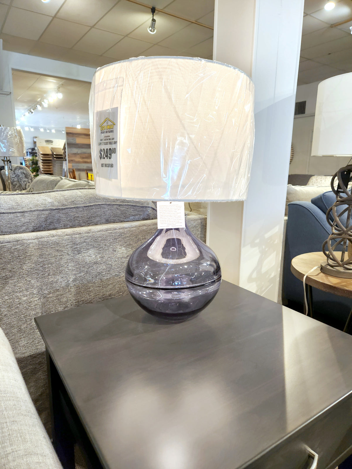 The Ashley L430784 Lemmitt Table Lamp with a blue glass base and white shade is on a dark side table in the store.
