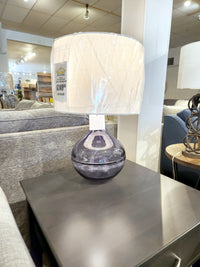 The Ashley L430784 Lemmitt Table Lamp with a blue glass base and white shade is on a dark side table in the store.