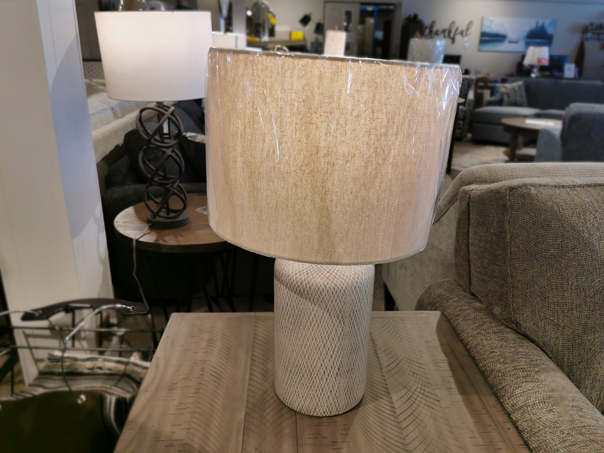 The Ashley L117964 Linus Table Lamp sits on a wooden table in a furniture store.