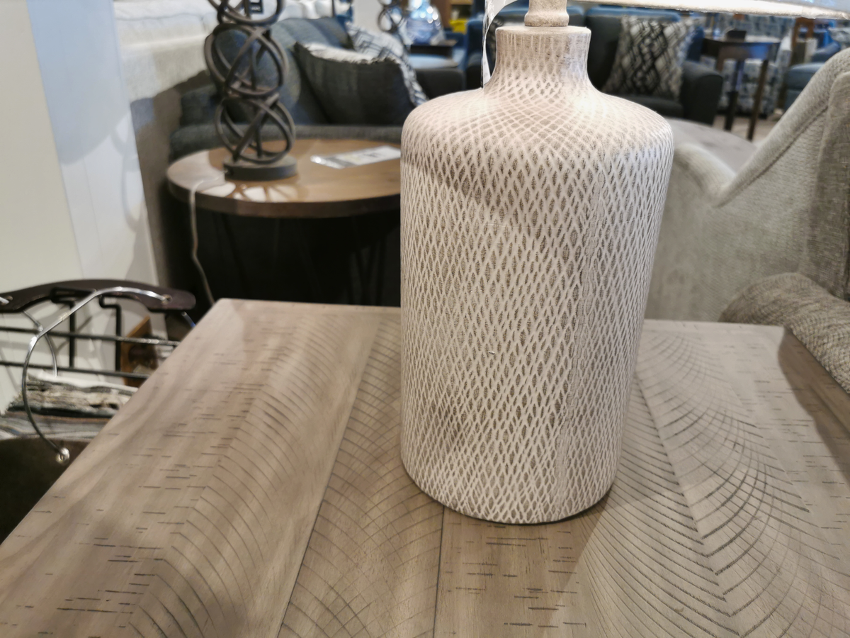 The Ashley L117964 Linus Table Lamp with an antique black finish sits on a wooden table in a showroom.