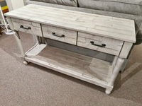 The Ashley Shawnalore Sofa Table with three drawers and a shelf is shown behind a gray sofa on carpeted flooring.