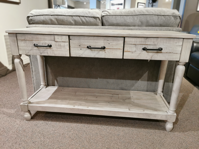 The Ashley Shawnalore Sofa Table with 3 drawers and a shelf sits behind a gray upholstered sofa.