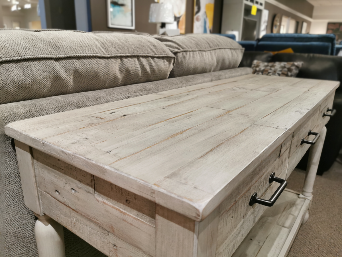 An Ashley Shawnalore Sofa Table in light pine with metal handles is displayed before a beige fabric sofa in-store.