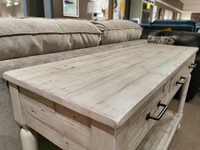 An Ashley Shawnalore Sofa Table in light pine with metal handles is displayed before a beige fabric sofa in-store.