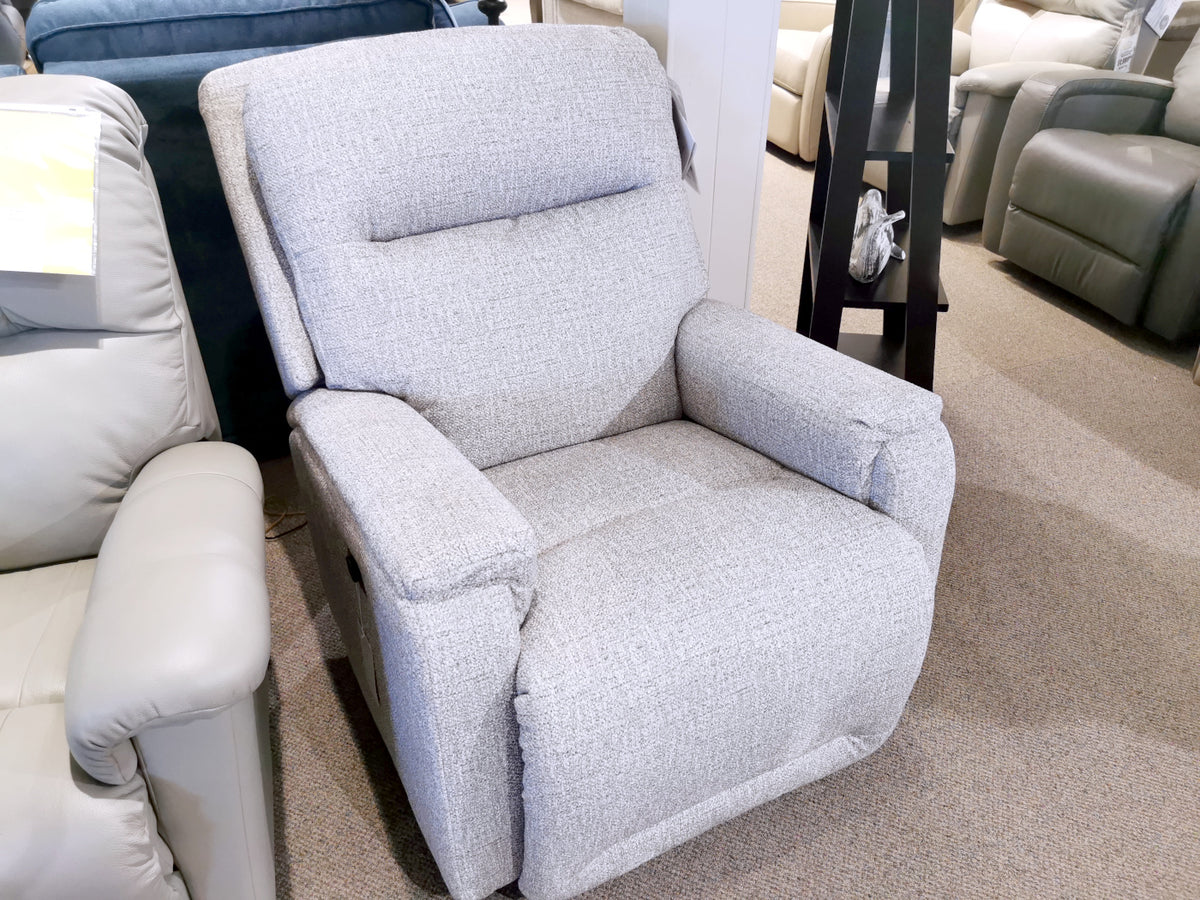 The 6NZ57 Derek Power Chair by Best, a gray upholstered recliner designed for ultimate comfort, is showcased in a showroom alongside other chairs on a beige carpeted floor.