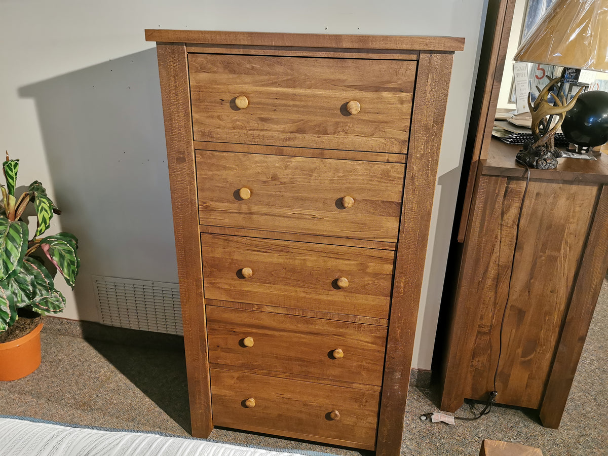 A Vokes 250 Rough Sawn 5-Drawer Chest stands tall beside a nightstand and a potted plant.