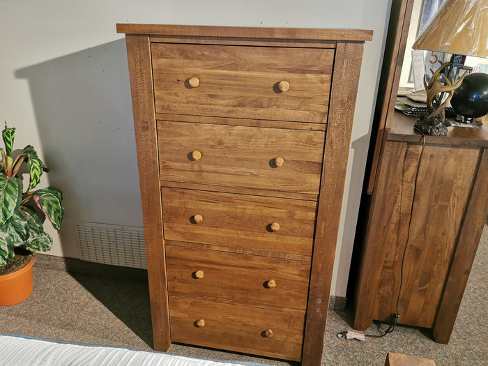 A Vokes 250 Rough Sawn 5-Drawer Chest stands tall beside a nightstand and a potted plant.
