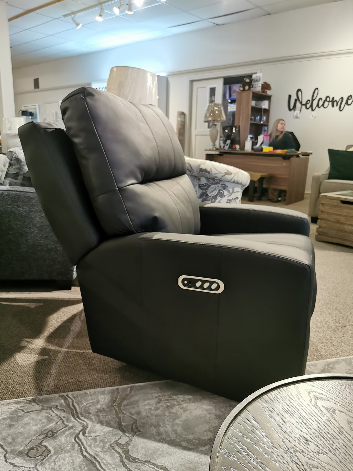 In a furniture showroom, the M38505 Power Reclining Loveseat by Decor-rest exudes elegance with its sleek black leather finish and powered features. From a side angle, a salesperson can be spotted at their desk in the background. The loveseat's headrest and lumbar support ensure unparalleled comfort for potential buyers.