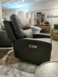 In a furniture showroom, the M38505 Power Reclining Loveseat by Decor-rest exudes elegance with its sleek black leather finish and powered features. From a side angle, a salesperson can be spotted at their desk in the background. The loveseat's headrest and lumbar support ensure unparalleled comfort for potential buyers.