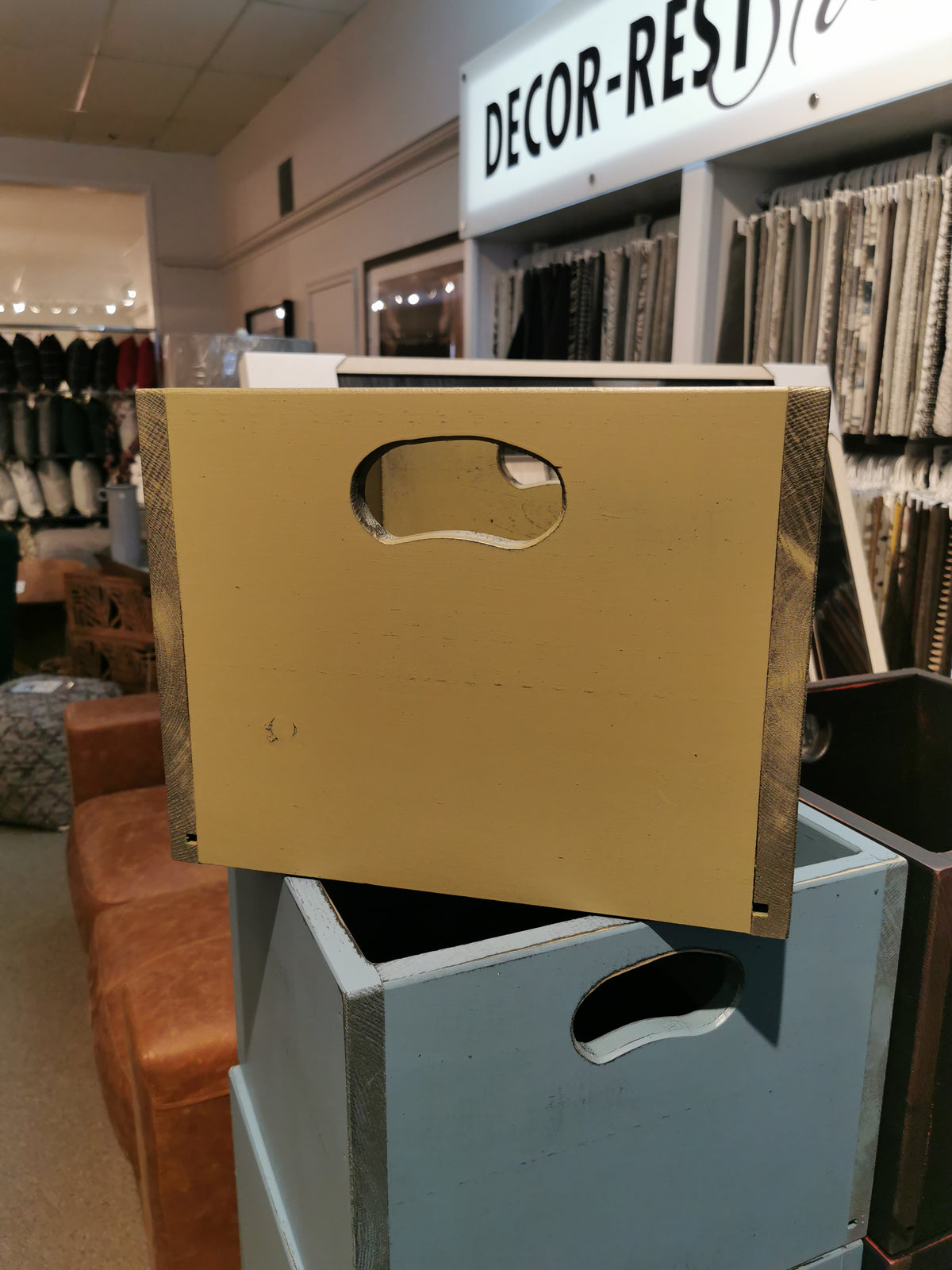 A yellow SB101 Record Crate by Mercana sits on a blue box with fabric samples and a "DECOR-REST" sign.