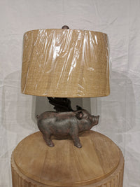 The Hogs Fly Table Lamp by Crestview Collection has a pig base and beige shade on a rustic wooden table.
