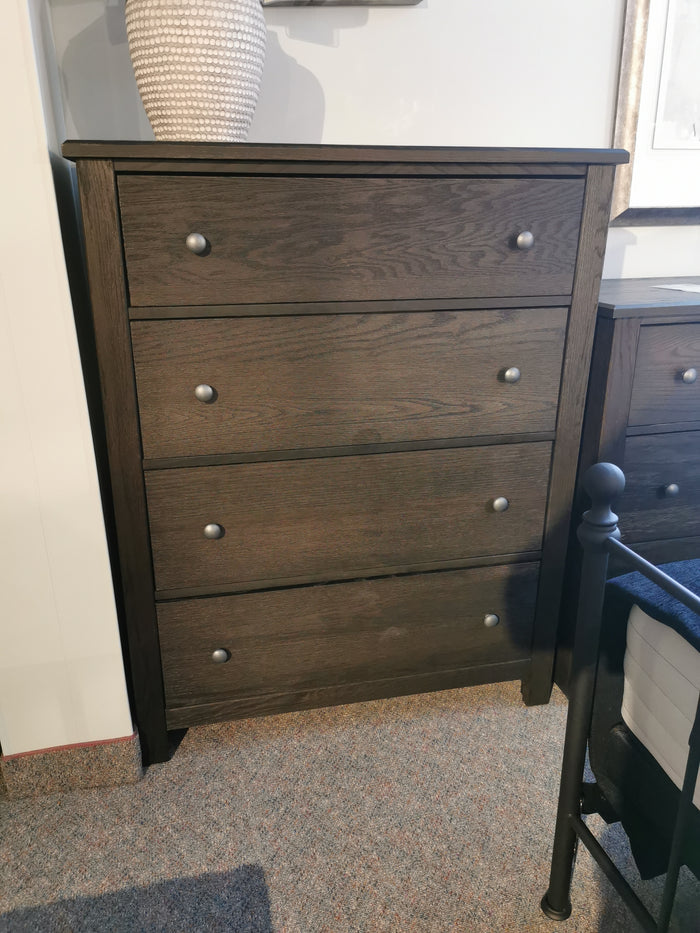 The Vaughan Bassett 10-114 Fundamentals Chest features drawers with metal knobs, set by a bed on carpet, topped by a woven vase.