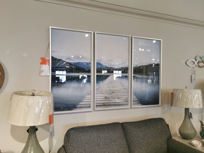 The GL549 Spring Dock Framed Art by Picture Depot, measuring 22" x 42" x 3", elegantly displays a scene of a wooden dock leading to a mountain lake. It complements a gray sofa, flanked by two lamps on either side.