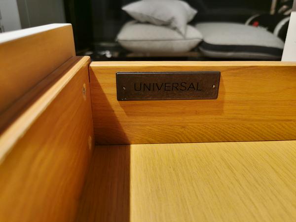 A wooden drawer from the Universal 987350 Summerhill 3 Drawer Night Table, "UNIVERSAL" label inside, cushions behind.