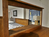 Wooden mirror reflecting a bedroom with a bed, lamp, artwork, and a Vokes 250 Rough Sawn 9-Drawer Dresser.