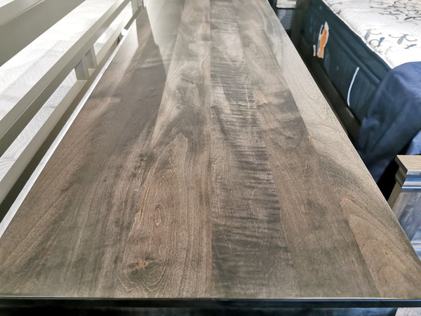 A close-up of the Vokes 462 Bayshore 7-Drawer Dresser’s wood grain tabletop beside a bed in a showroom.