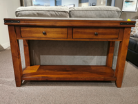 The Mango Sofa Table by Winners Only, with two drawers and a shelf, is set against a light gray sofa in a carpeted room.
