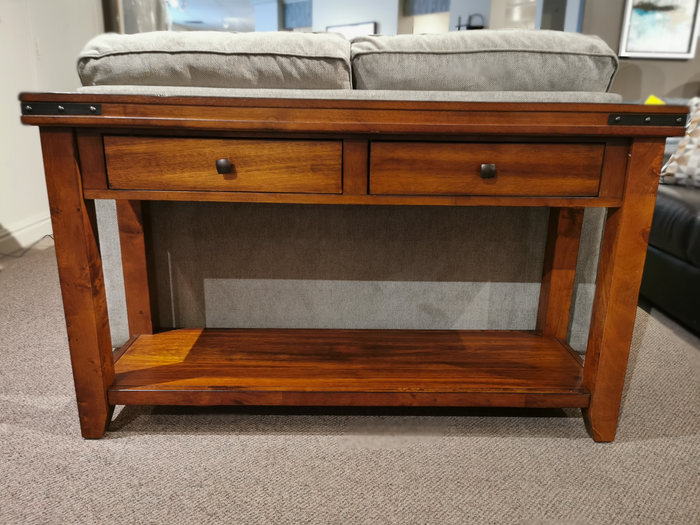 The Mango Sofa Table by Winners Only, with two drawers and a shelf, is set against a light gray sofa in a carpeted room.