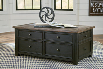 The T736 Tyler Creek Coffee Table by Ashley, featuring four drawers and a wheel, sits on a patterned rug with an open book.