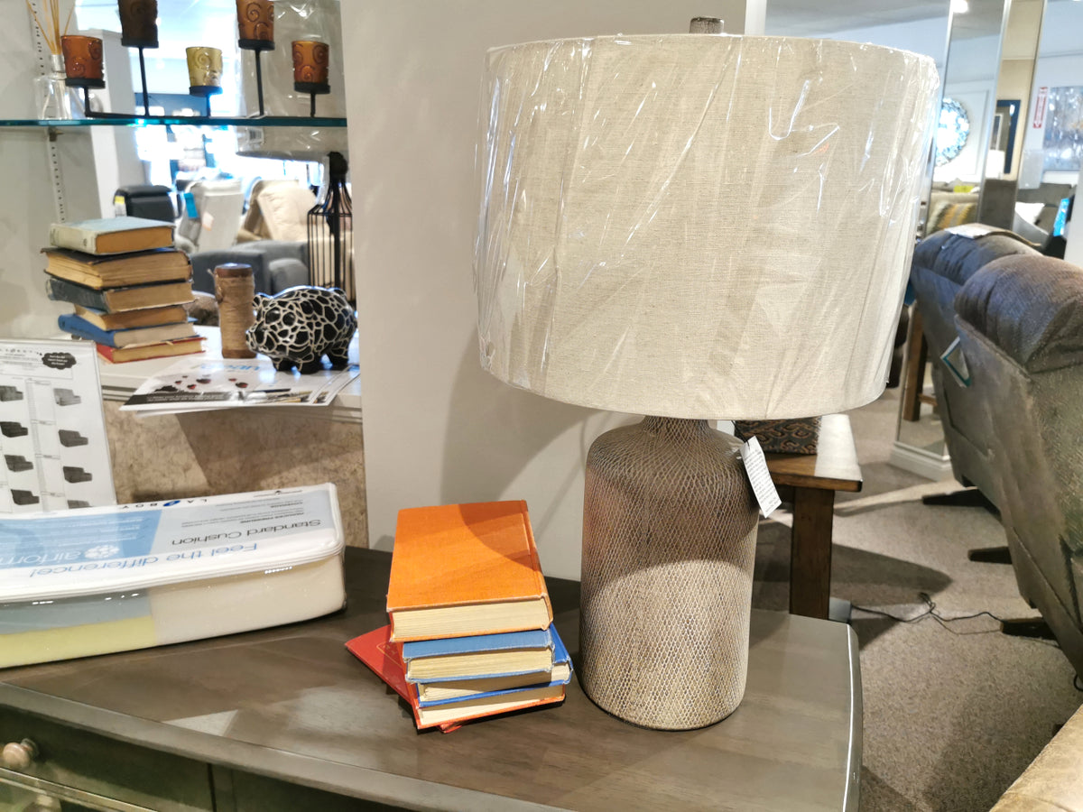 An Ashley L117964 Linus Table Lamp with ceramic base and plastic-wrapped shade sits on a wooden console in-store.