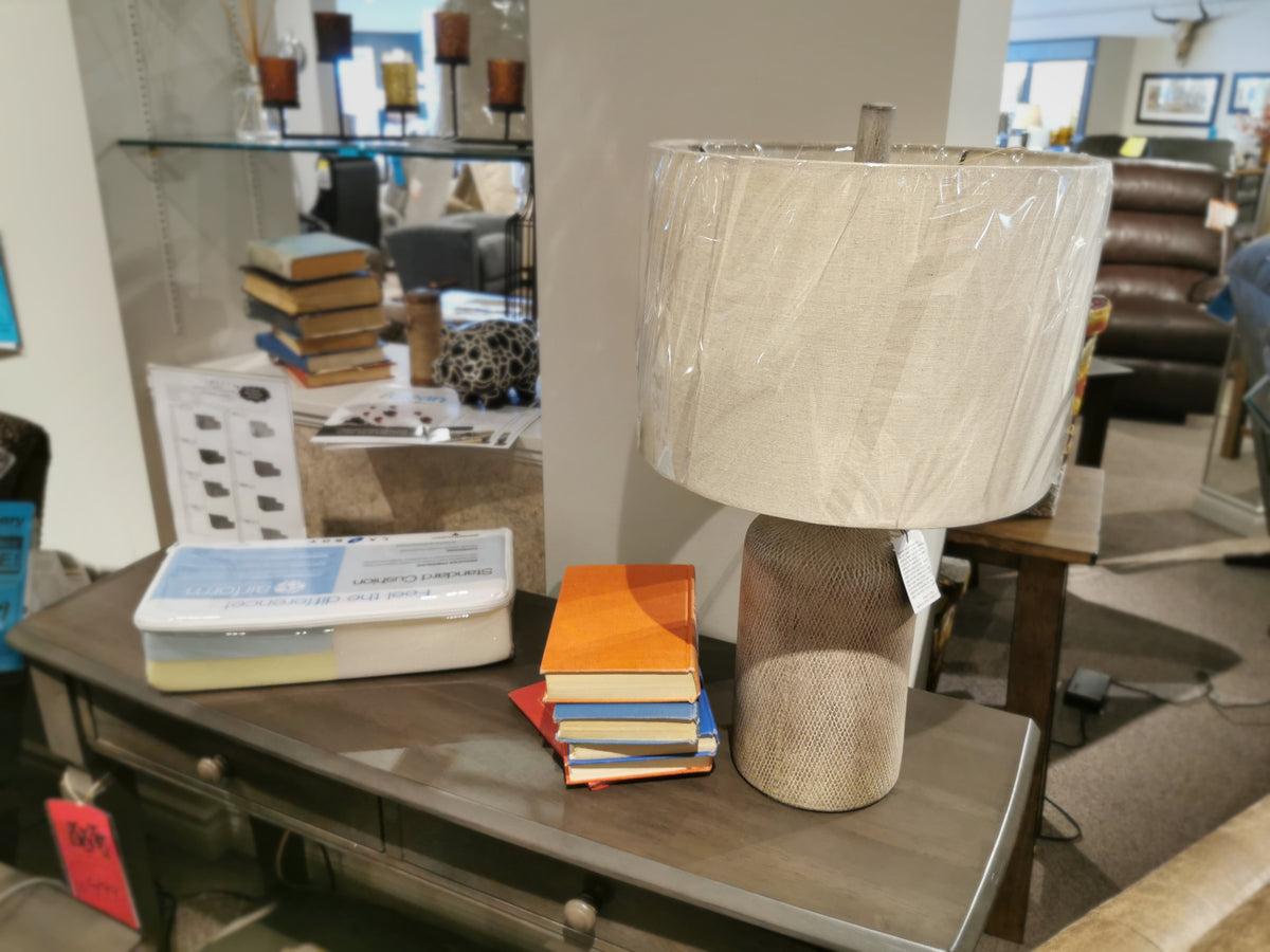 Ashley L117964 Linus Table Lamp with ceramic base and plastic-wrapped shade displayed on a wooden store console.