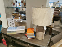 Ashley L117964 Linus Table Lamp with ceramic base and plastic-wrapped shade displayed on a wooden store console.