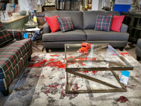 Modern setup with gray sofa, plaid chairs, and the Ashley Coylin Coffee Table featuring an angled metal base and glass top.