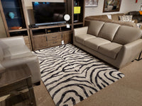 A living room display with a beige sofa set, wooden TV stand, and Kalora Sable 9652/P384 zebra pattern rug on carpet.