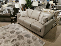 The Oxford Sofa by Leather Craft in gray leather with two patterned pillows sits beside a rug and side table in the showroom.