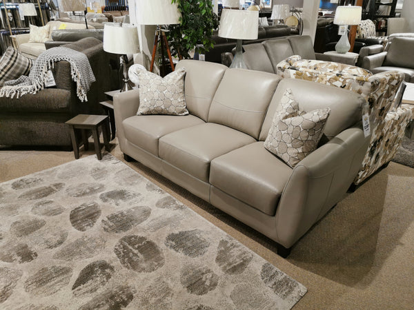 The Oxford Sofa by Leather Craft in gray leather with two patterned pillows sits beside a rug and side table in the showroom.